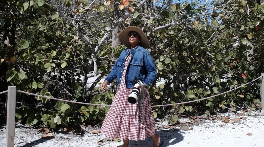 Woman in a straw hat and denim jacket holding a camera, walking on a sandy path by sea grape bushes and palm trees under a clear blue sky.