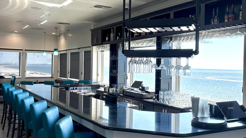 bar photo with blue barstools and black bar with windows around overlooking the beach and natural light shining through