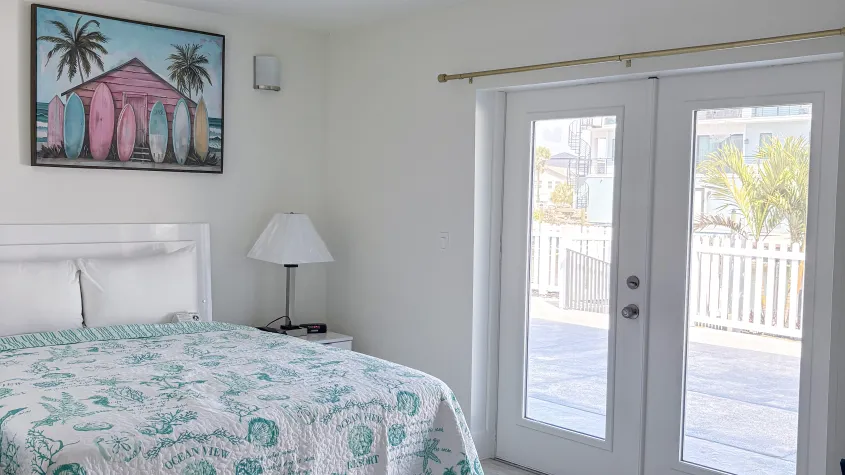 bedroom with bed, pillows, green and white sheets, picture above bed, glass double doors, natural sunlight, wood floors, lamp