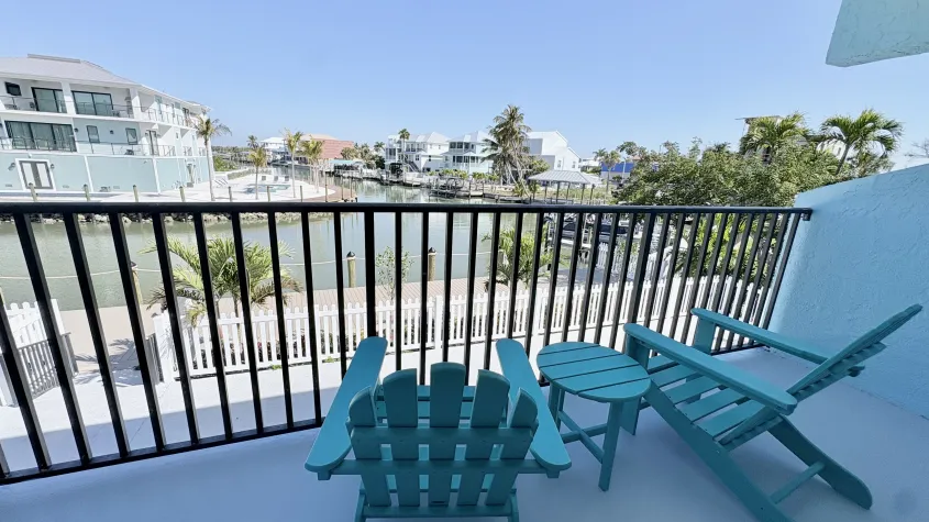 two blue lounge chairs and small table on a balcony, with black railing overlooking the back bay water boats palm trees