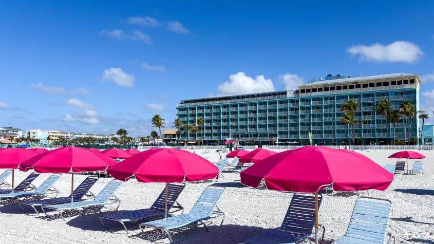 pink umbrellas and blue lounge chairs lined up on a beach with a blue hotel in the background and blue skiies