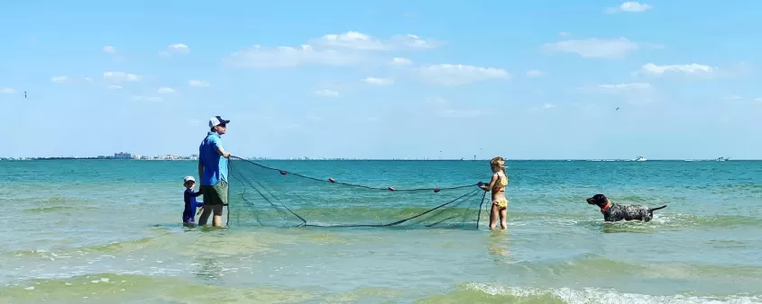 Two people throwing a cast net with dog nearby in the water.